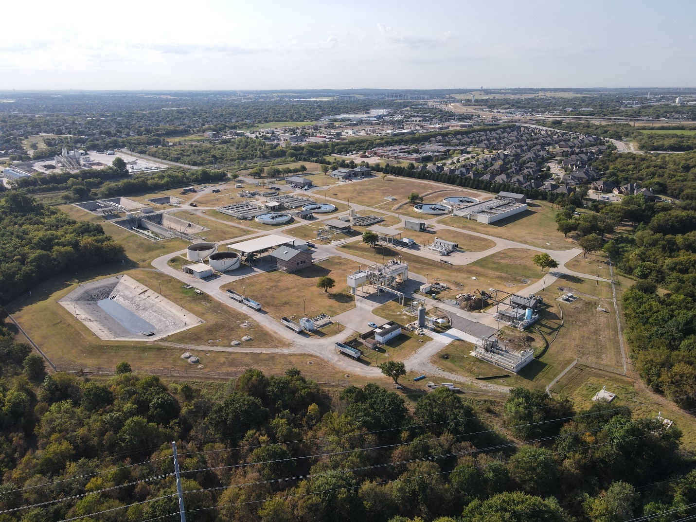 Aerial view of a wastewater treatment facility with circular tanks, buildings, and surrounding residential and wooded areas