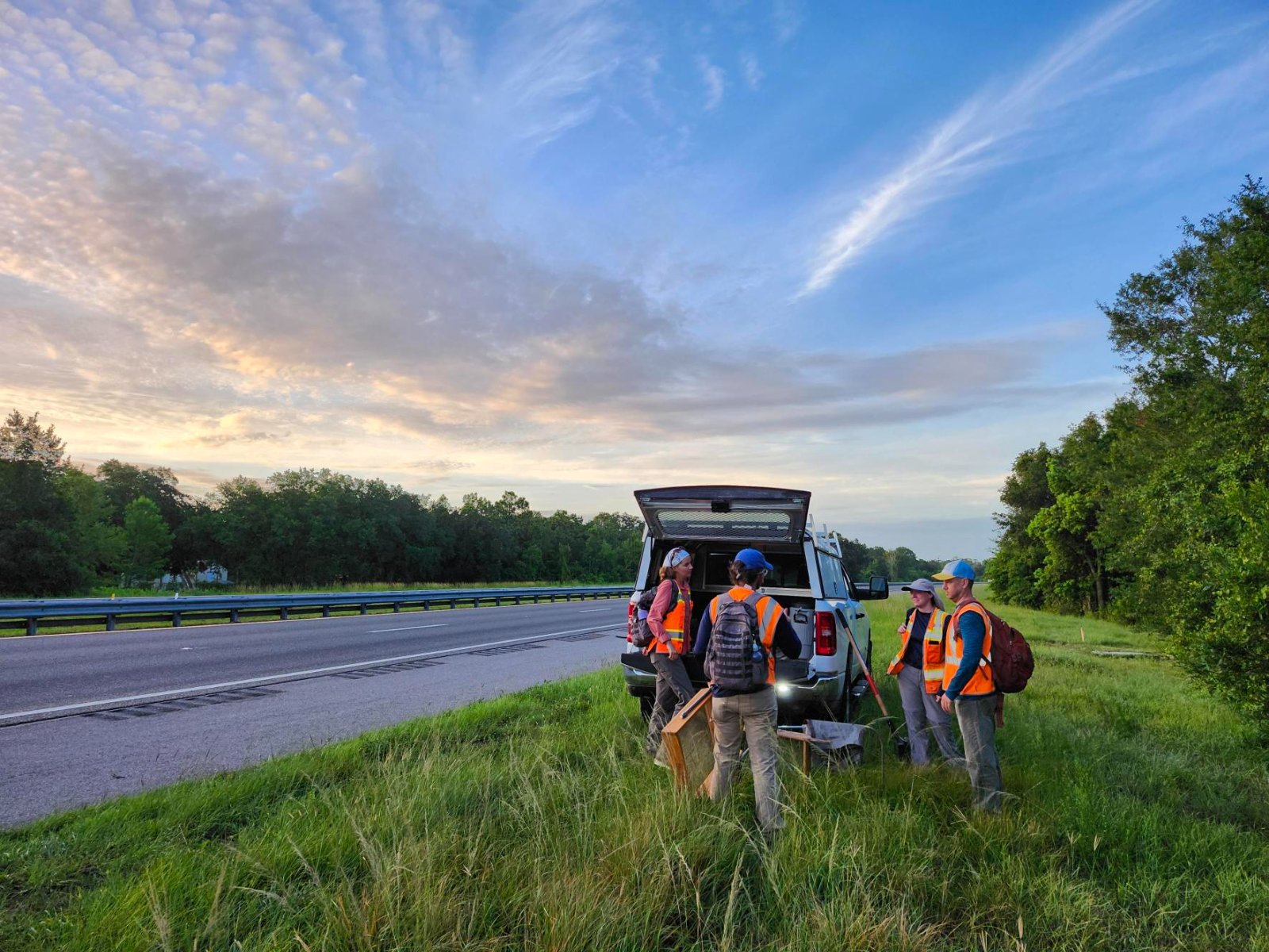 Group of workers in safety vests standing beside a pickup truck on grassy roadside next to a highway