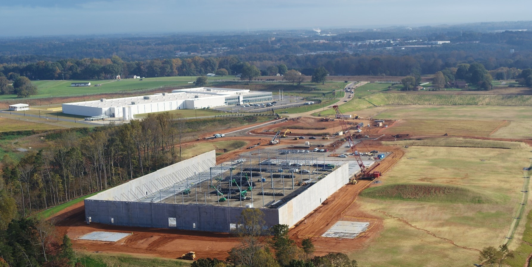 Large industrial building under construction with concrete walls, steel framing, cranes, and earthwork in a rural landscape