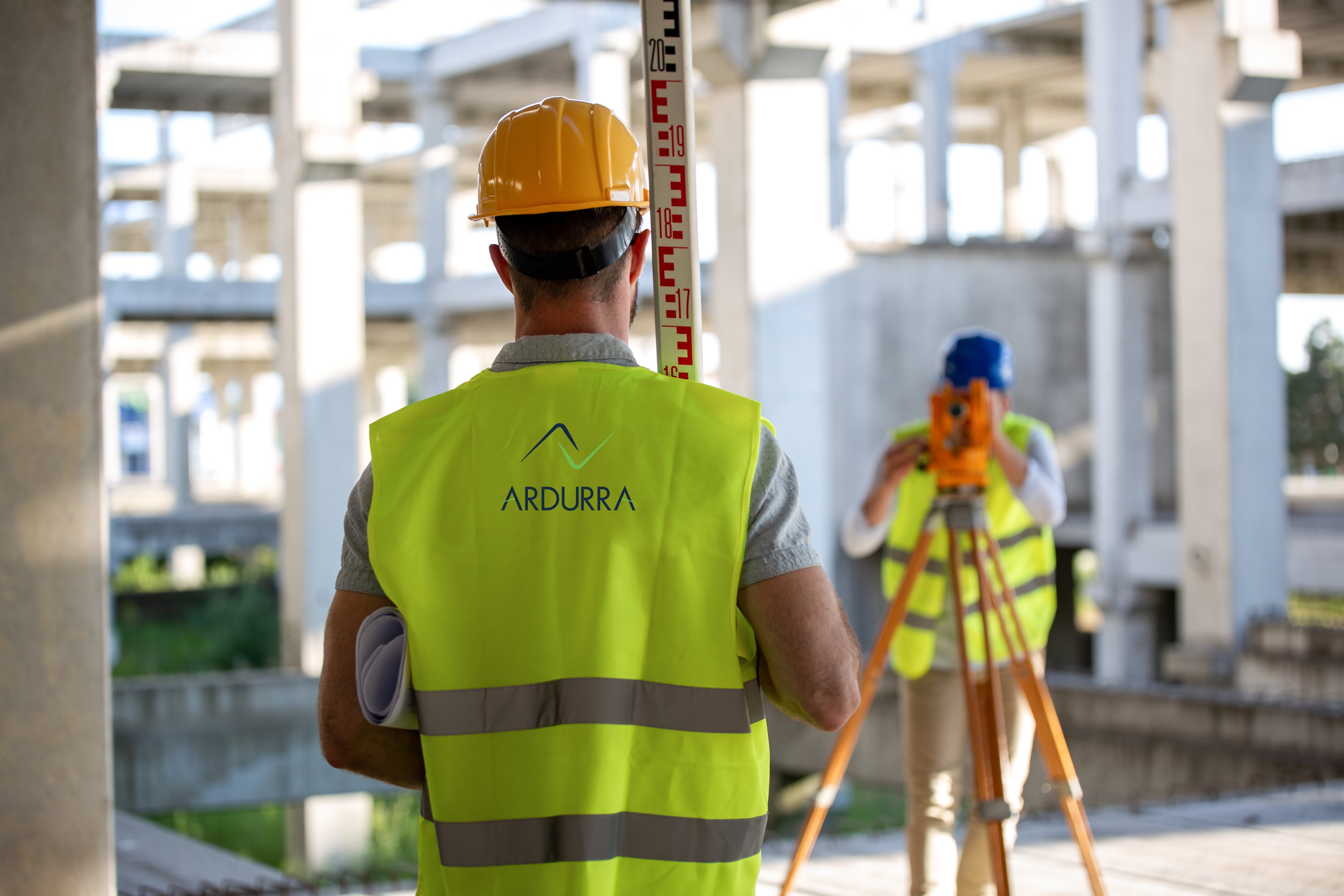 Construction worker in high-visibility vest holding measuring rod while another operates surveying equipment on a building site